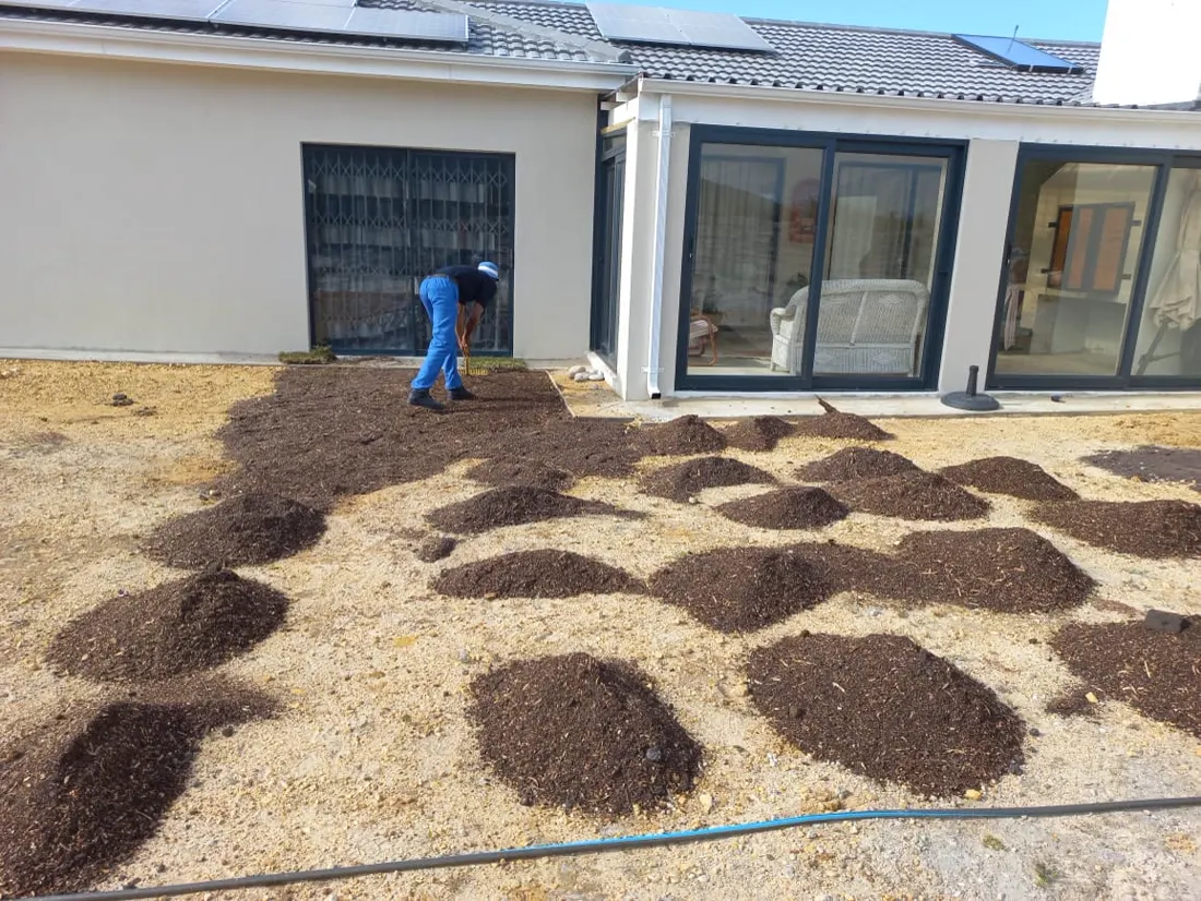 A gardener spreading soil across bare ground beside a house, preparing it for new lawn